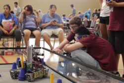 Two students wearing maroon shirts and jeans crouch outside of a FIRST Tech Challenge playing field as they discuss the calibration of their robot, pictured on the left. 