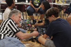 A volunteer wearing a black and white striped referee's shirt works closely with a student at a computer. They are in the process of fixing an error in a FIRST Tech Challenge-style robot, partially pictured in the background. 